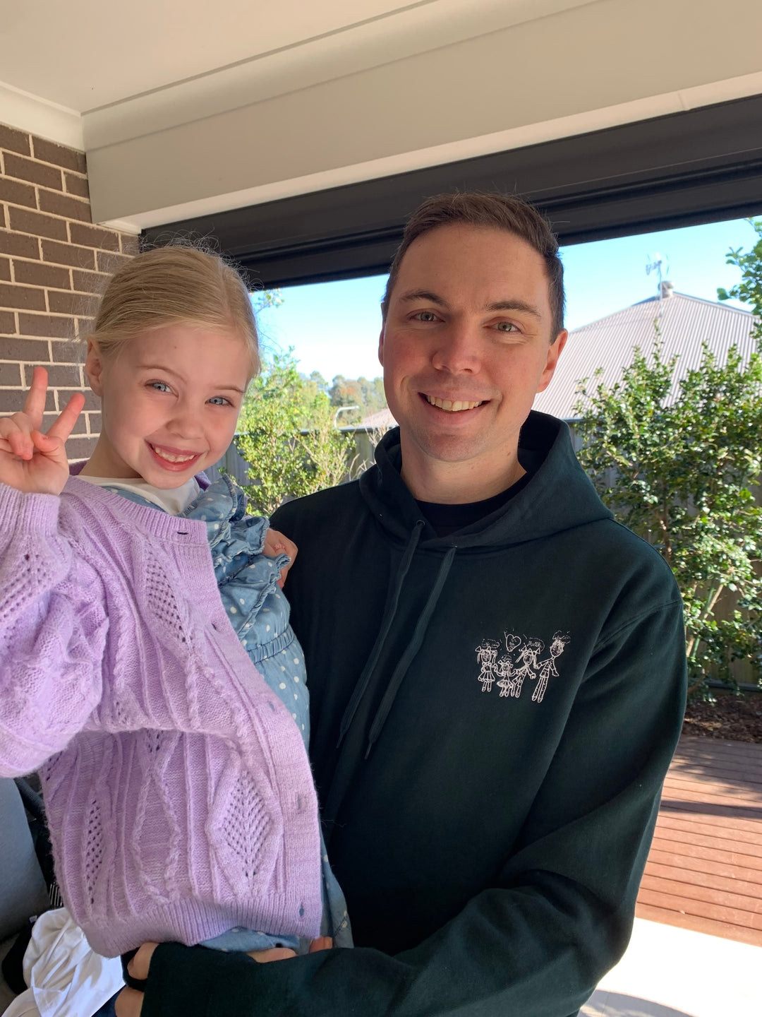 Dad holding his daughter, both smiling, family drawing embroidered on a black RK Threads hoodie
